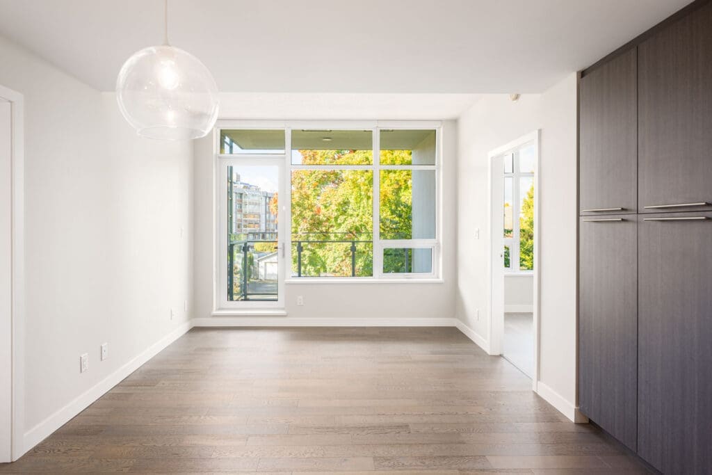 Bright, empty room with large windows, light wooden floors, white walls, a modern pendant light, and built-in dark cabinets; trees and buildings visible outside.