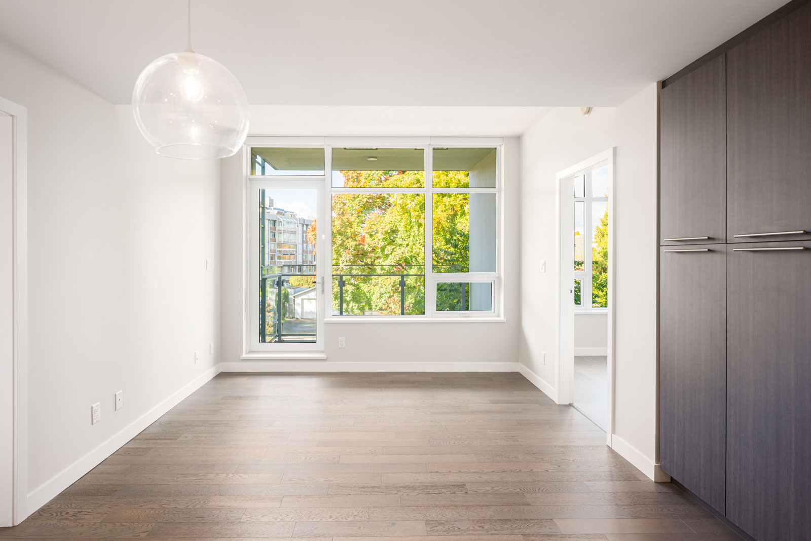Bright, empty room with large windows, light wooden floors, white walls, a modern pendant light, and built-in dark cabinets; trees and buildings visible outside.