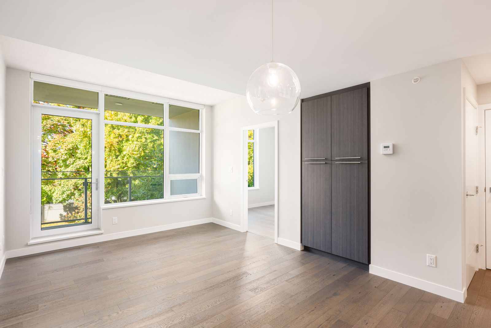 Bright empty room with large windows, light walls, dark wood floors, a hanging light fixture, and built-in dark cabinets, with trees visible outside.