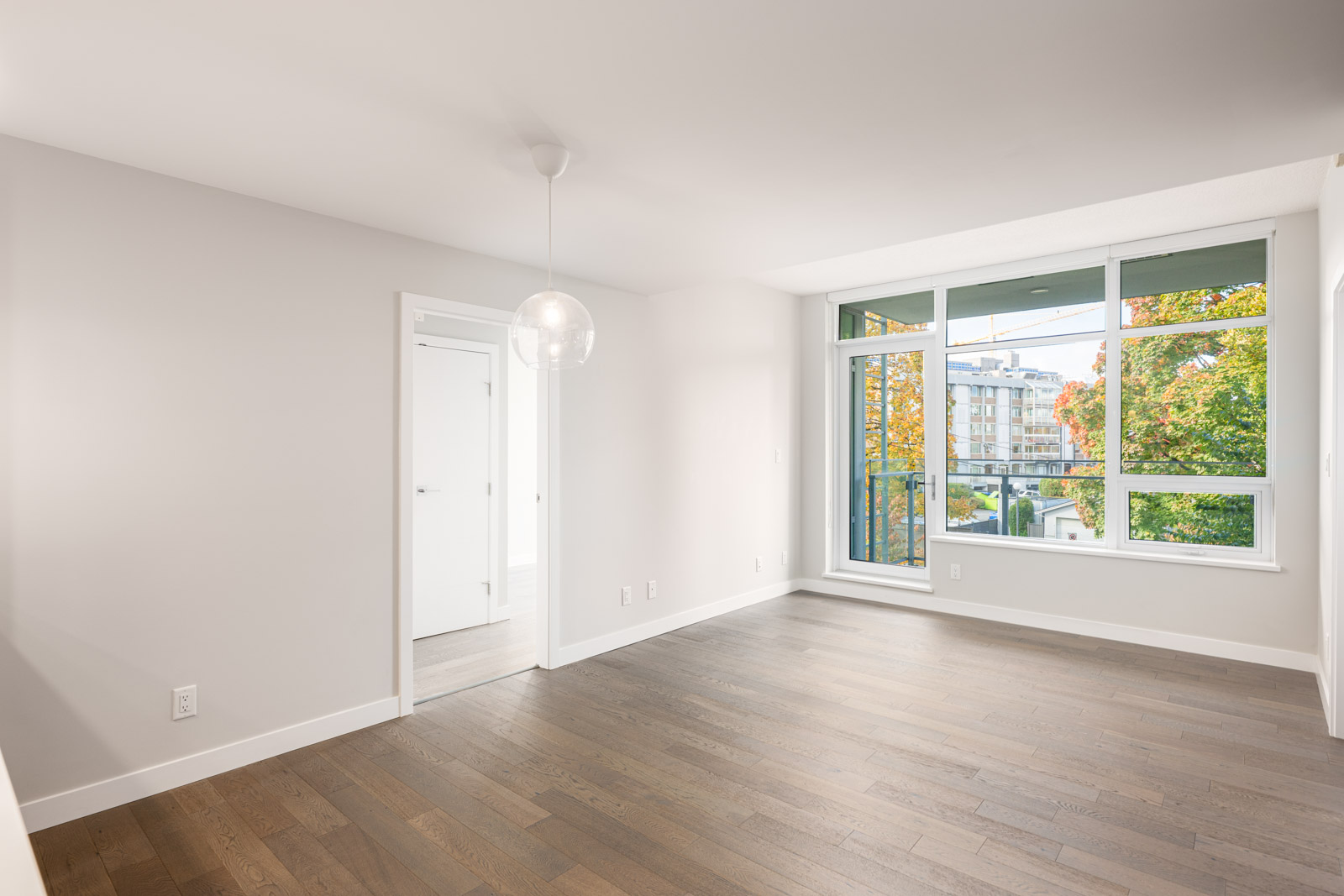 Empty, modern apartment room with light hardwood floors, white walls, a glass pendant light, large windows, and a door leading to another room; trees and buildings visible outside.