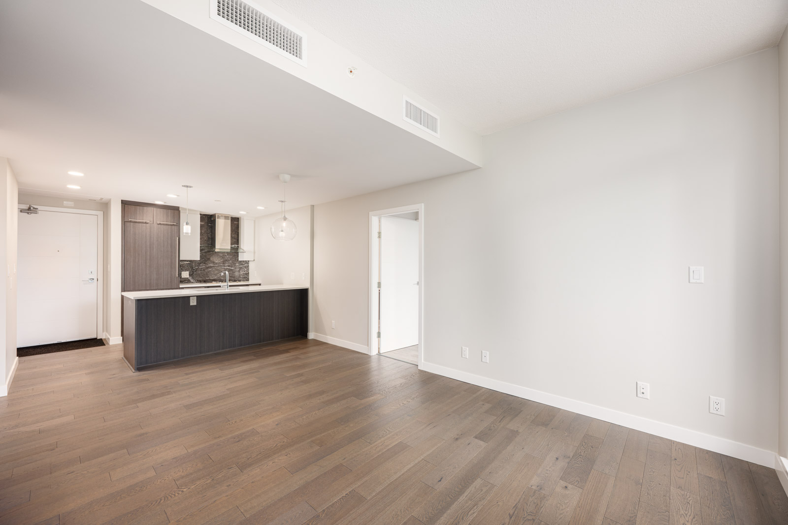 Unfurnished modern apartment interior with hardwood floors, white walls, open kitchen area, and a door leading to another room.