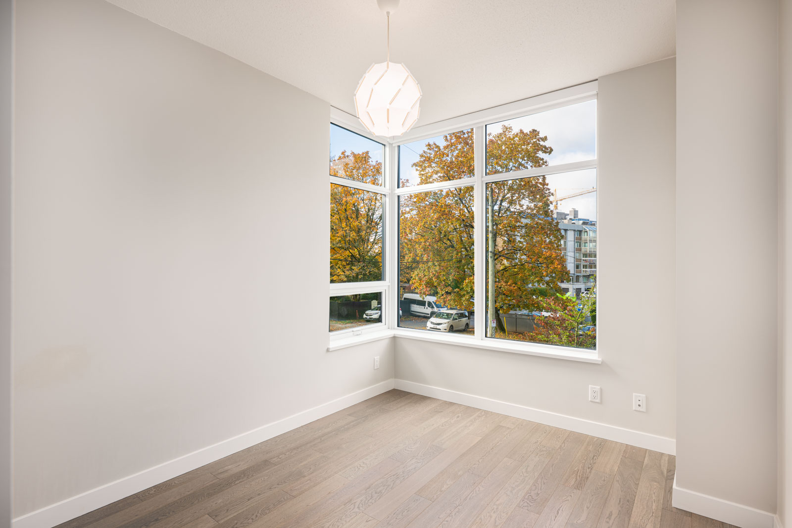 Empty corner room with light-colored walls, large windows showing autumn trees outside, modern light fixture on ceiling, and light wood flooring.
