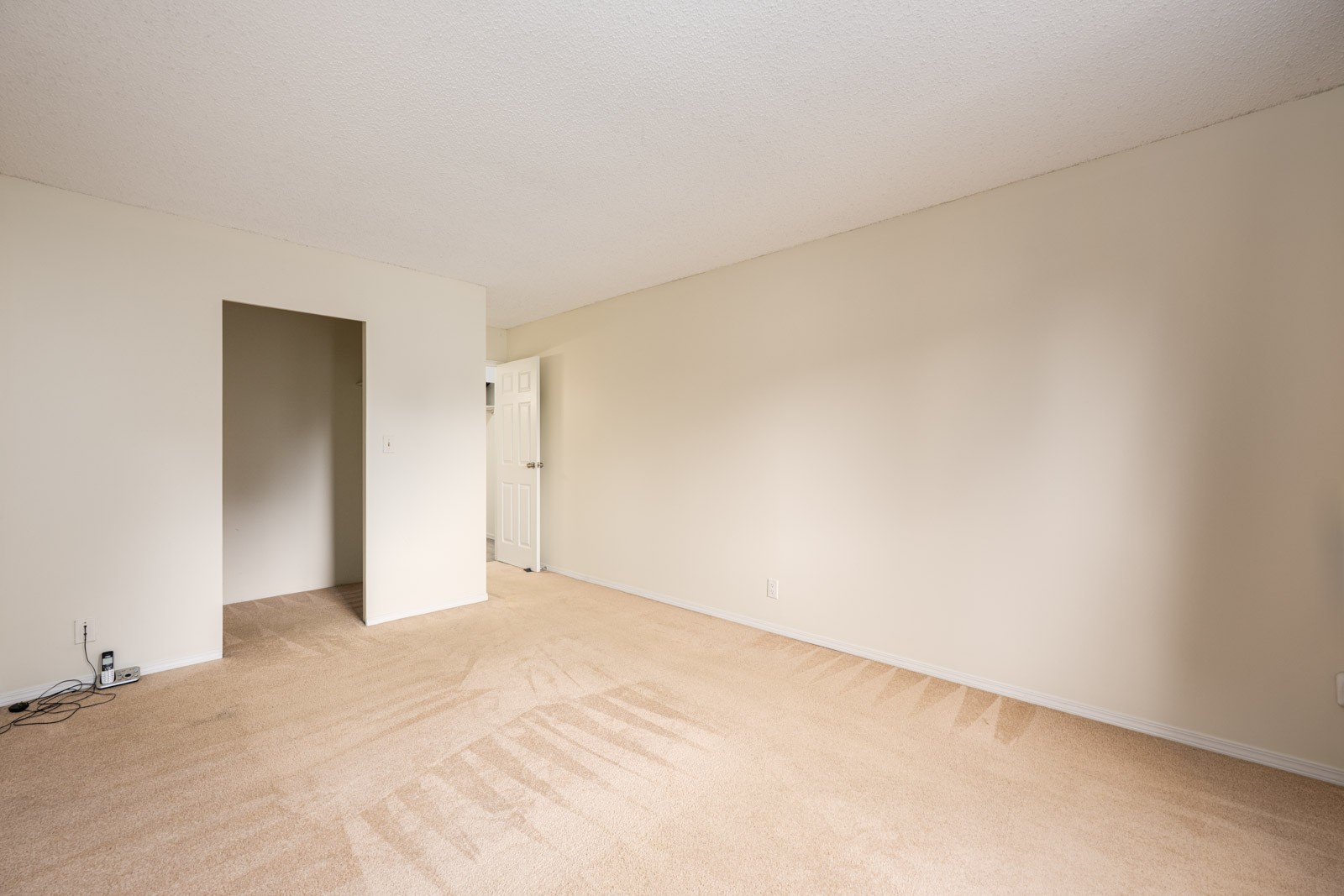 Empty room with beige carpet, off-white walls, open doorway, and a closed white door; visible vacuum lines and a few power outlets near the wall.