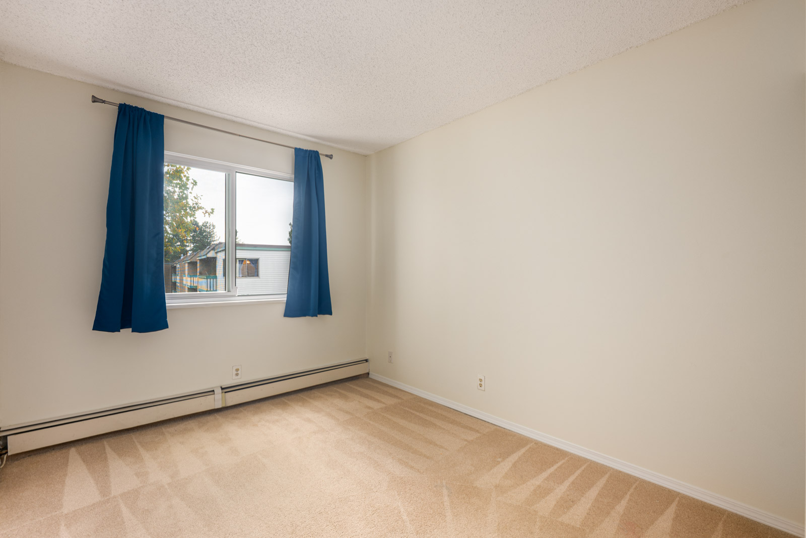 Empty room with beige carpet, white walls, a window with blue curtains, and baseboard heating beneath the window.