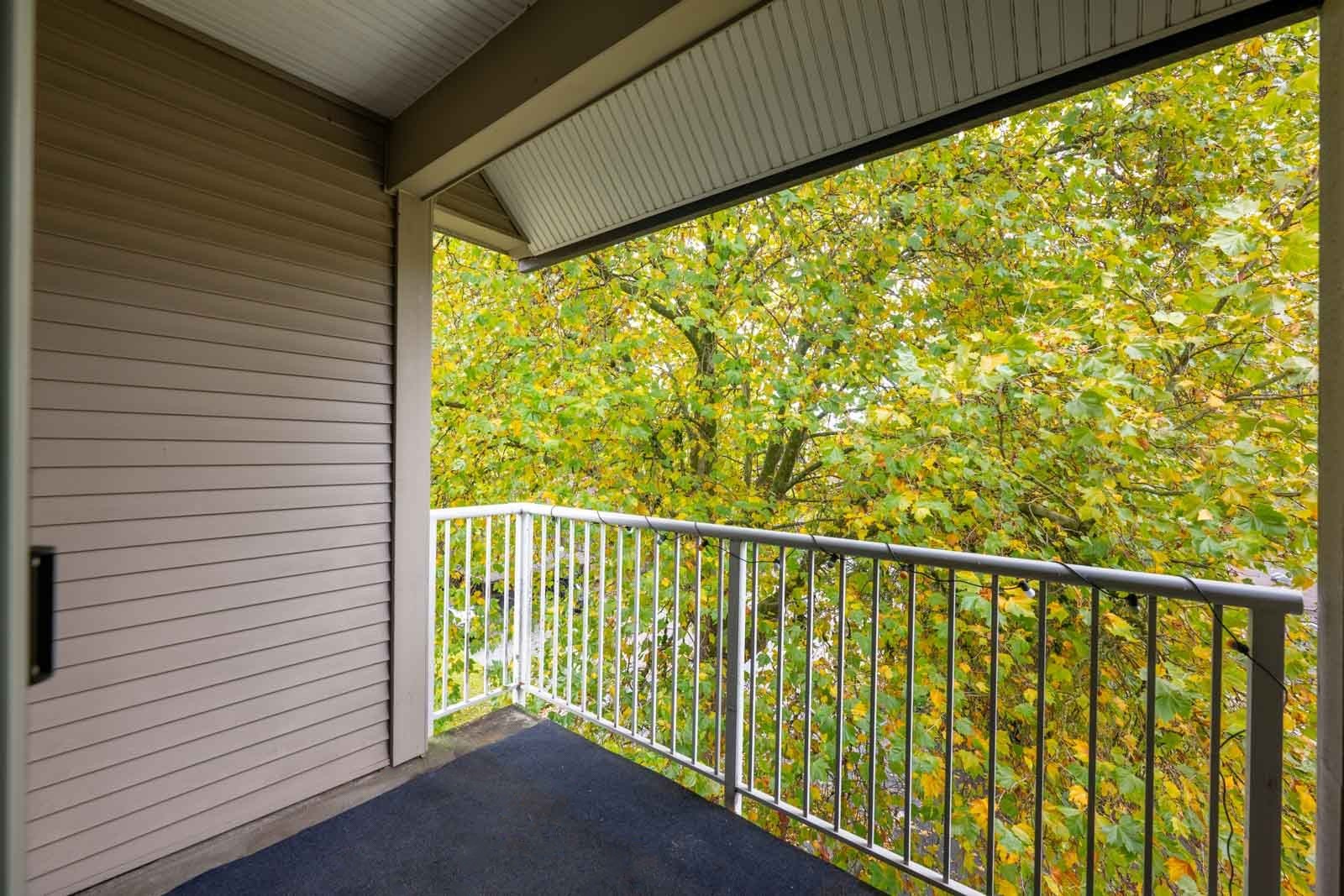 A small balcony with a white metal railing overlooks dense green and yellow-leaved trees, attached to a building with beige siding and a slanted roof.