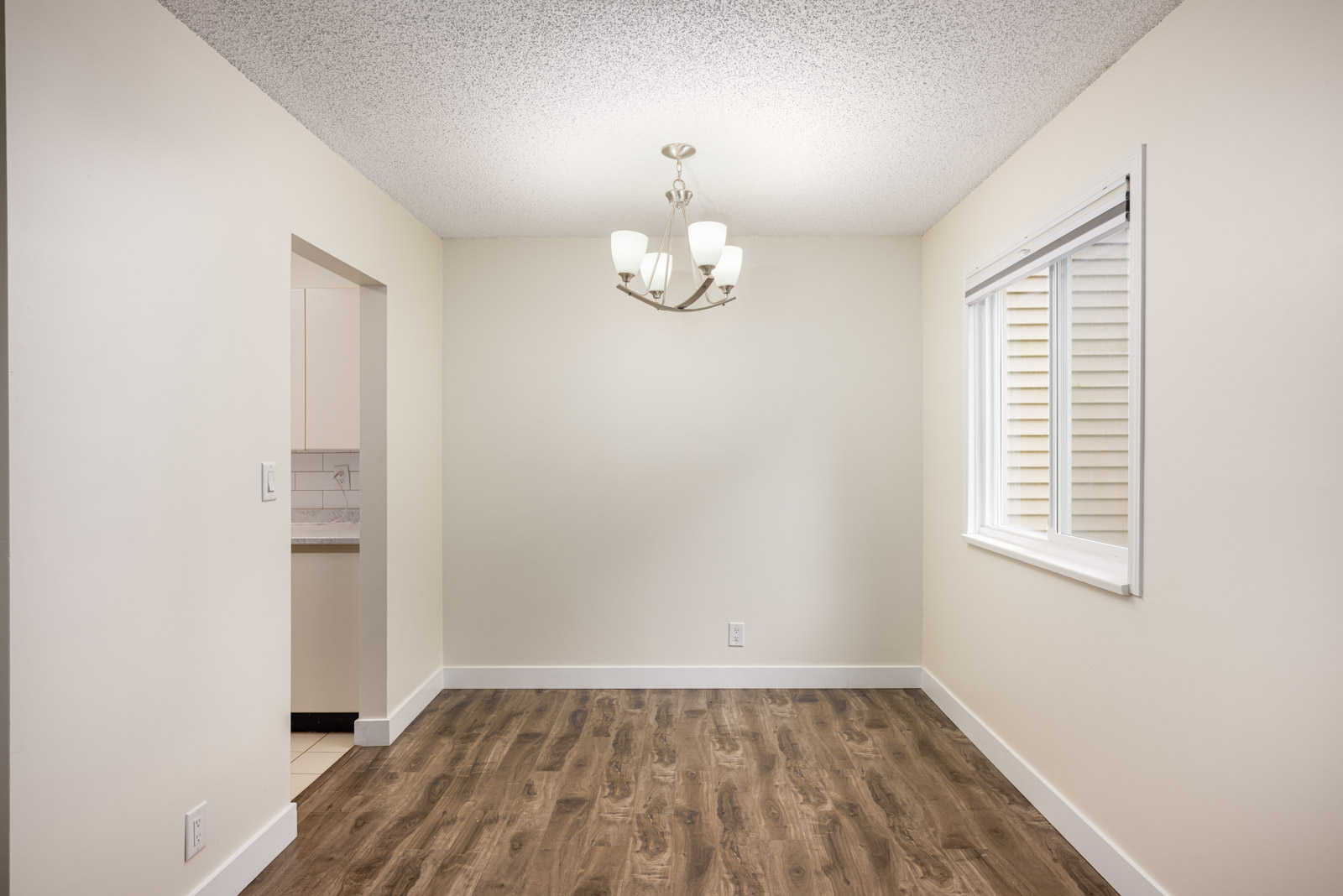 Empty dining room with wood flooring, white walls, a ceiling light fixture, and a window on the right side. An open doorway on the left leads to a kitchen area.