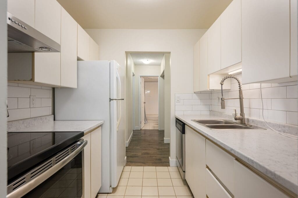 Narrow kitchen with white cabinets, stainless steel appliances, tiled backsplash, and a single sink, leading to a hallway with wood flooring.