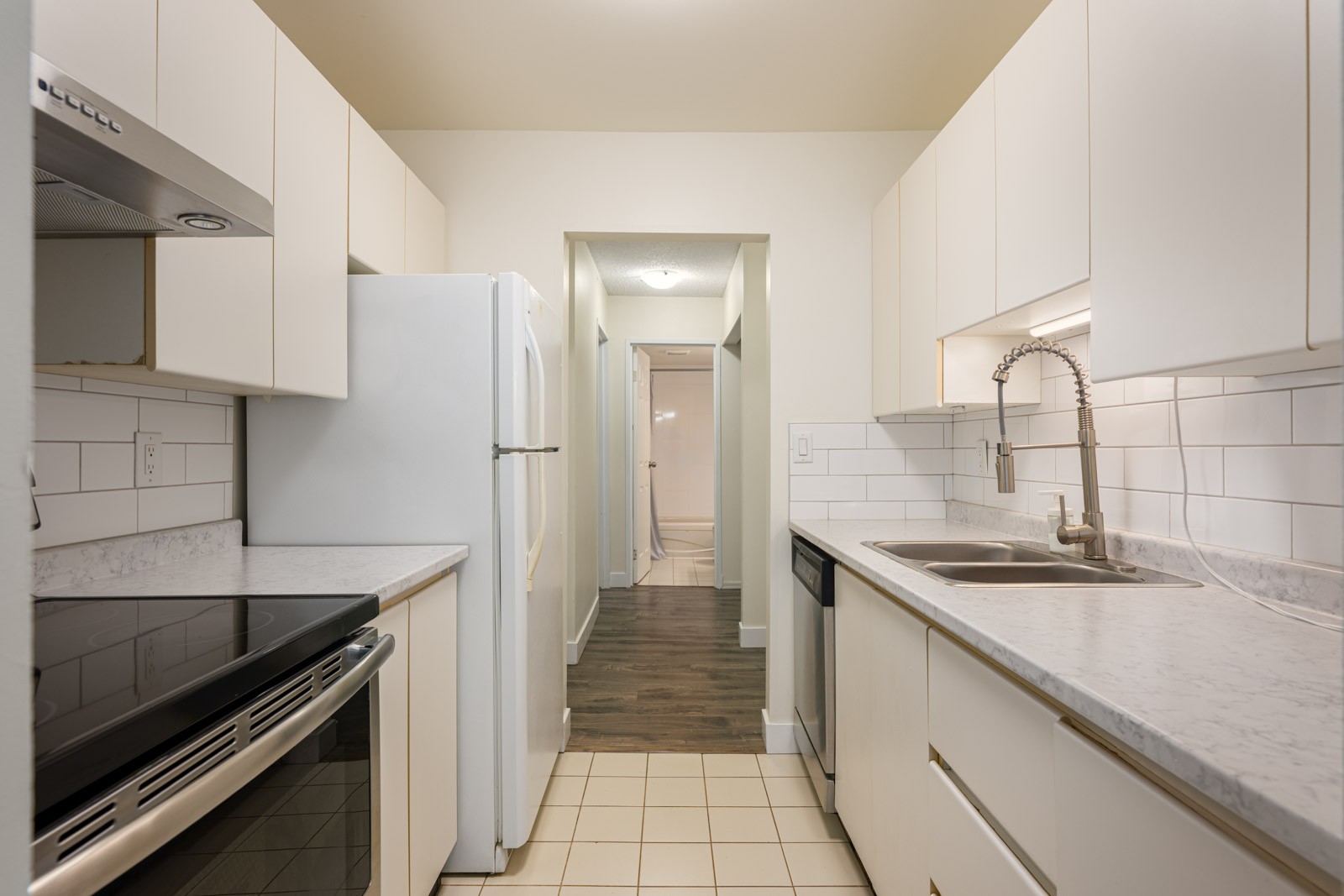 Narrow kitchen with white cabinets, stainless steel appliances, tiled backsplash, and a single sink, leading to a hallway with wood flooring.