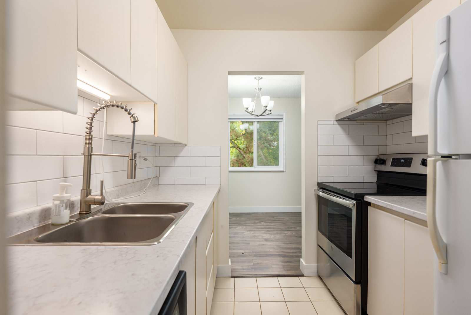 Modern galley kitchen with white cabinets, marble countertops, stainless steel sink, electric stove, and a view into a dining area with large window and chandelier.