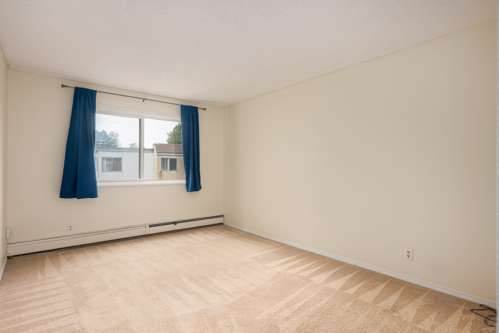 Empty carpeted room with cream-colored walls, a window with blue curtains, and baseboard heating along the wall below the window.