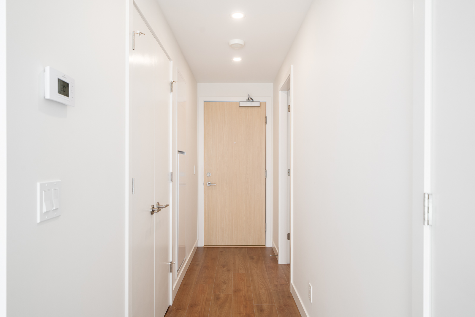 A bright, narrow hallway with white walls, wooden floor, closed white doors on each side, and a light wood entry door at the end.
