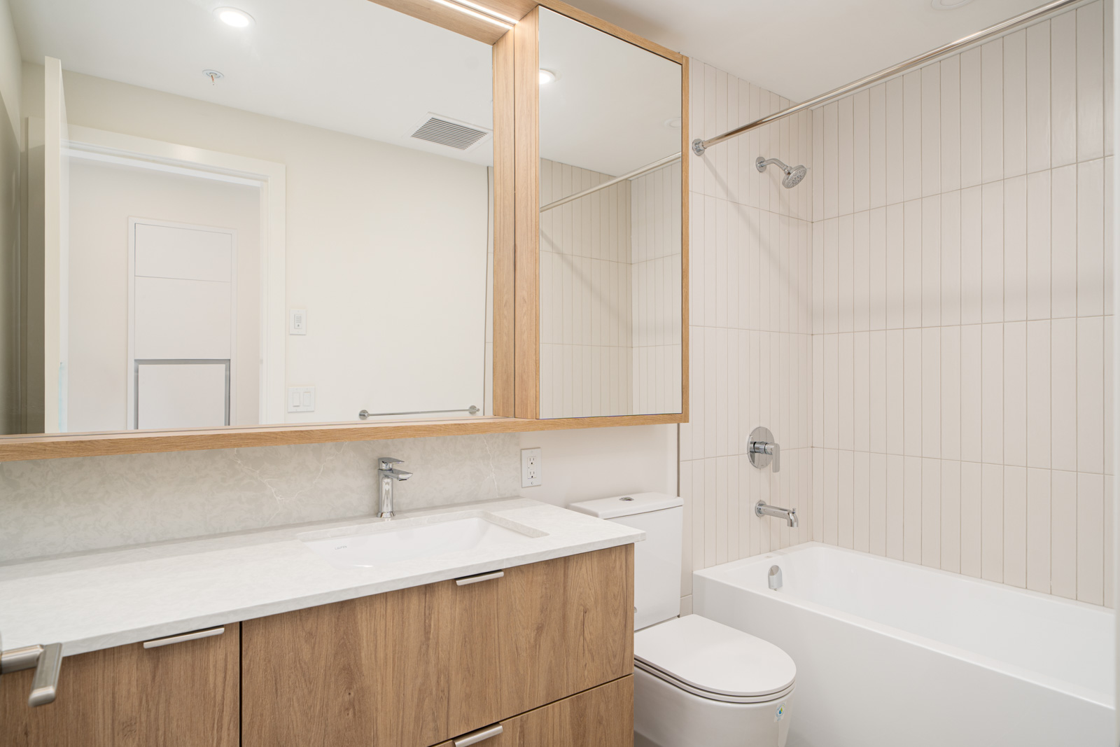 Modern bathroom with wood vanity, large mirror, white countertop, toilet, and a bathtub with overhead shower; light tiled walls and neutral color scheme.