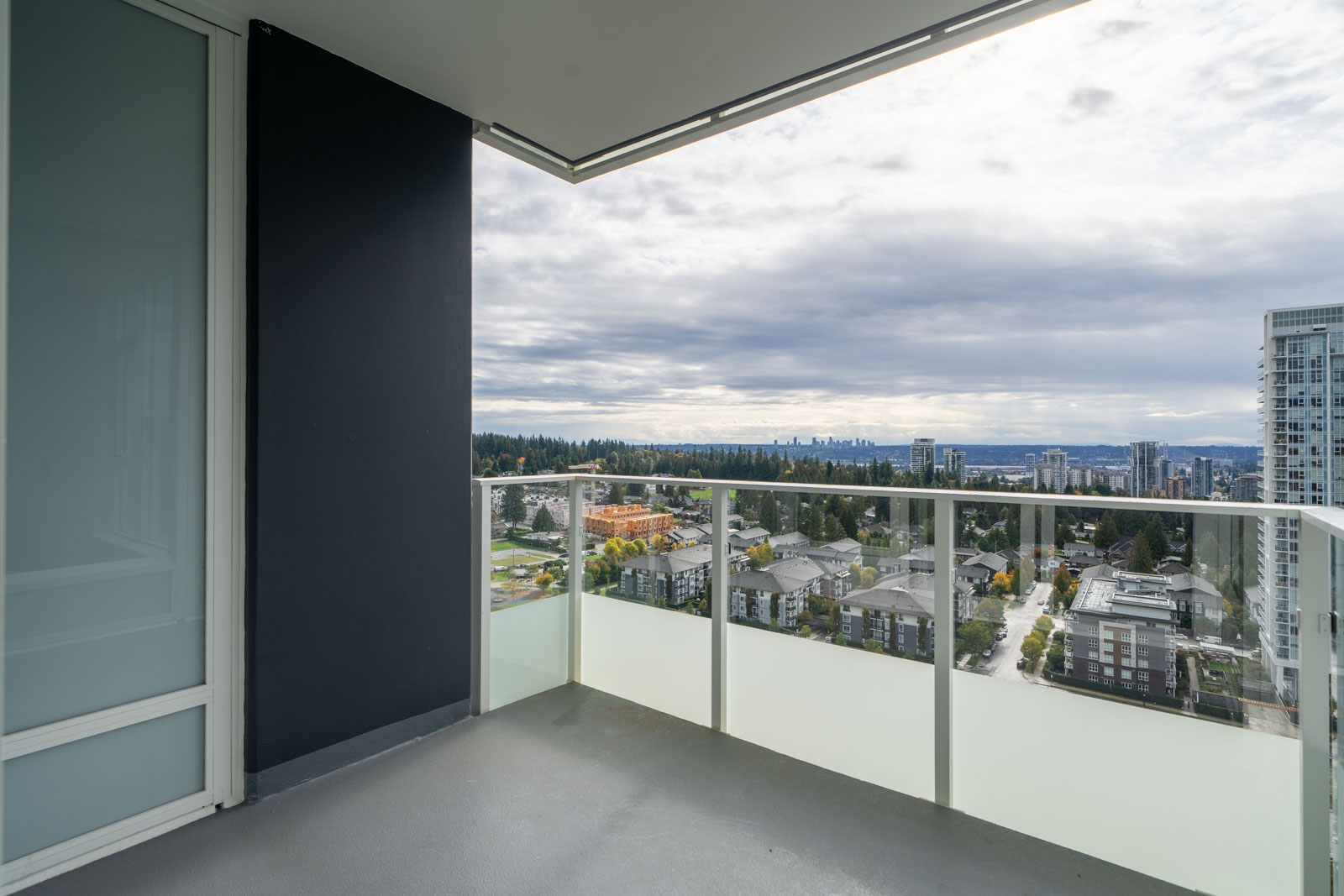 A high-rise balcony with glass railing overlooks a cityscape with buildings, trees, and a cloudy sky in the distance.