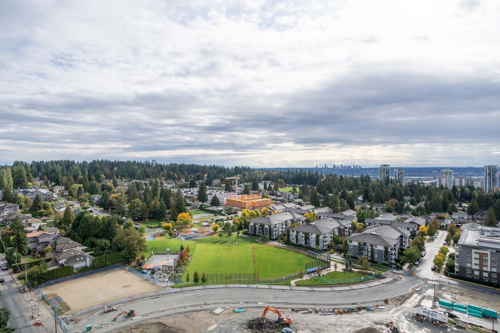 Aerial view of a suburban neighborhood with apartment buildings, a construction site, a sports field, and trees under a cloudy sky.