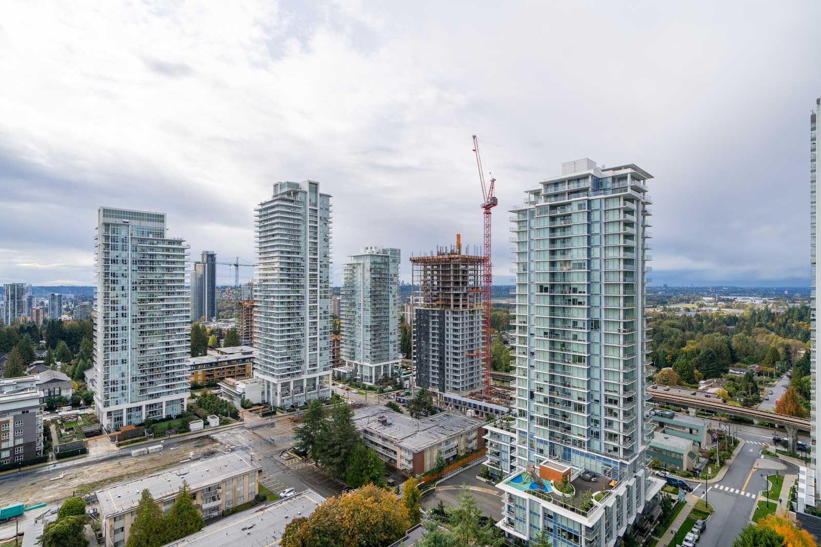 A cityscape with several modern high-rise buildings, a construction crane, and a mix of greenery and roads under a cloudy sky.