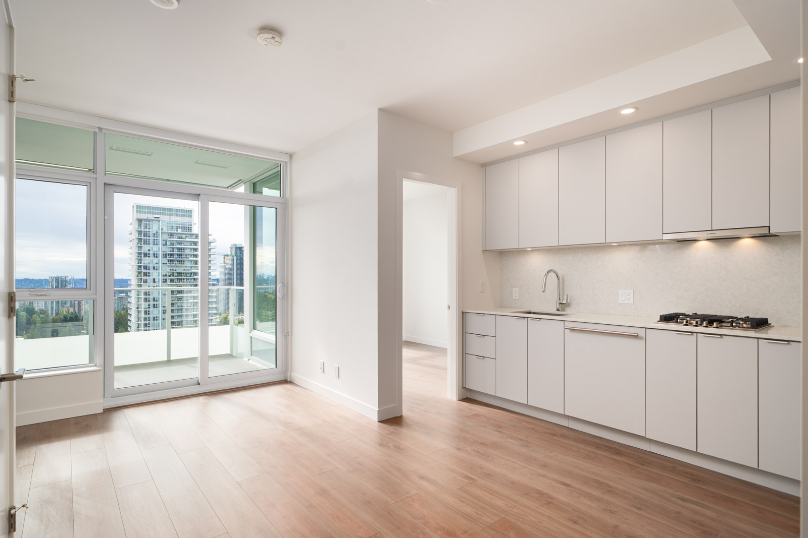 Modern, unfurnished apartment with light wood flooring, white kitchen cabinets, built-in stove, and large windows leading to a balcony with a city view.