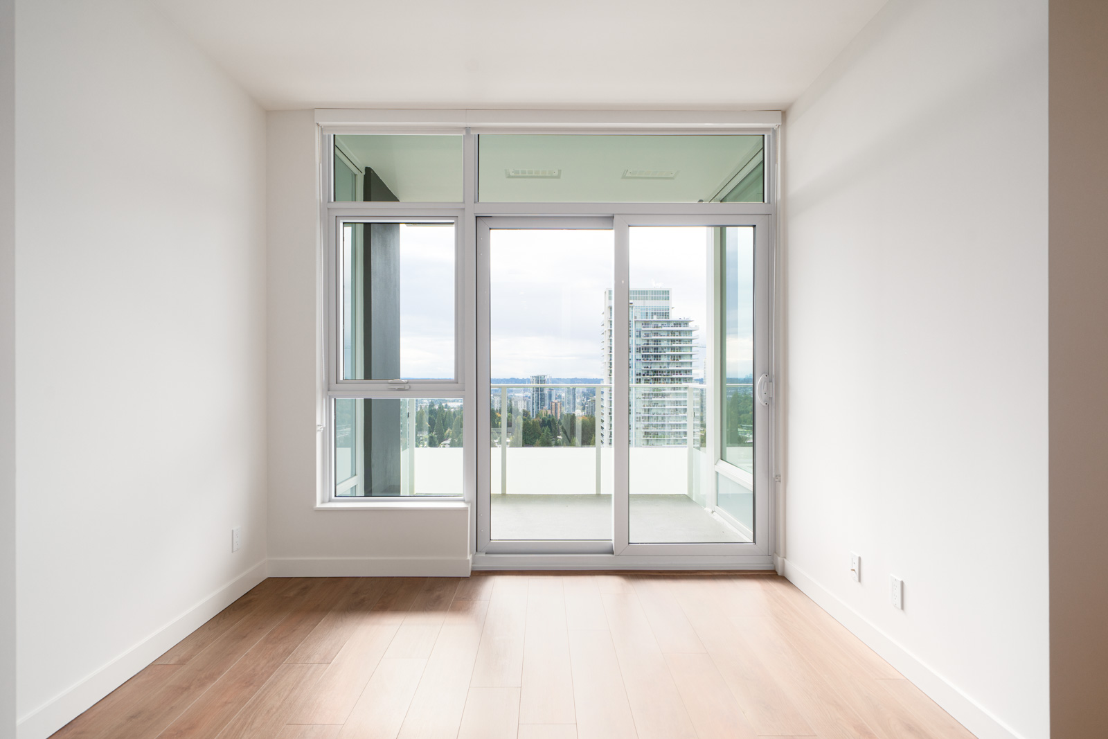 Empty room with light wood flooring, white walls, and large sliding glass doors leading to a balcony with a cityscape view.