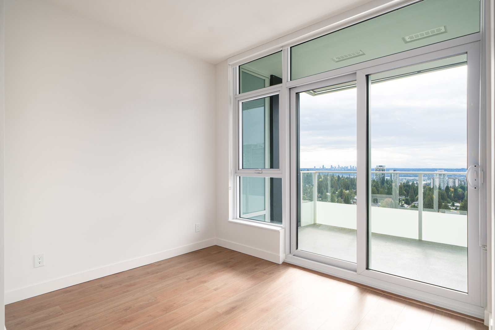 Empty room with light wood flooring, white walls, and large sliding glass doors leading to a balcony with a city skyline view.