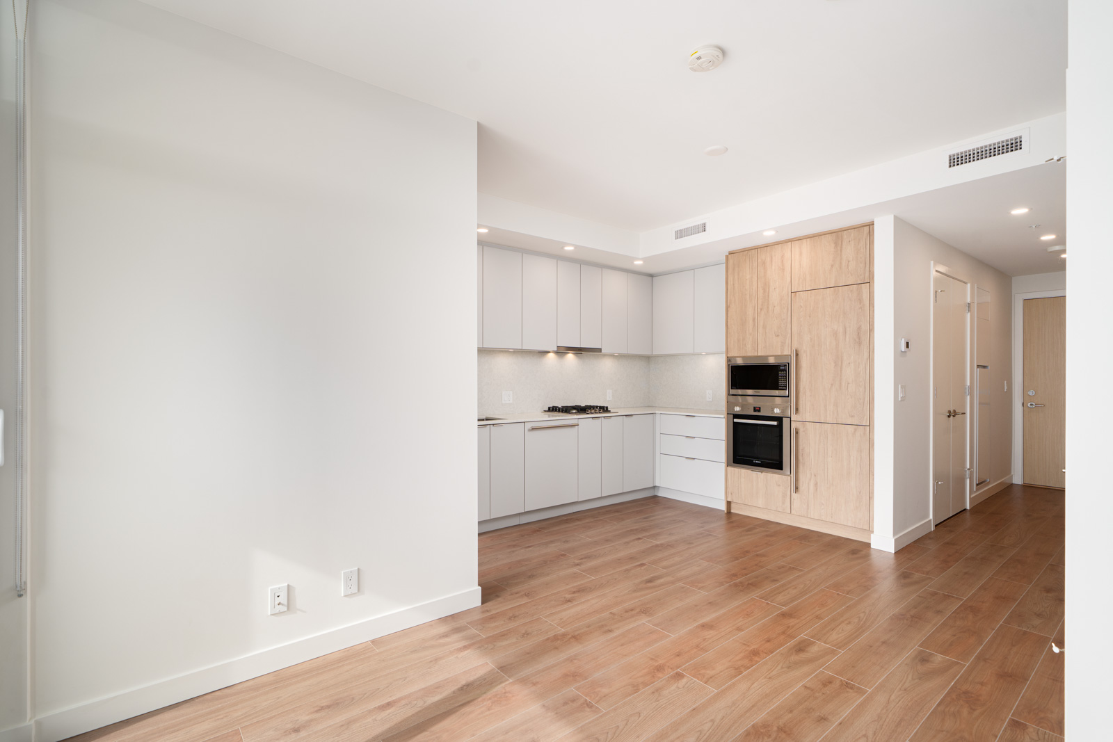 Modern, unfurnished kitchen and living area with light wood flooring, built-in appliances, white cabinets, and neutral walls in a new apartment.