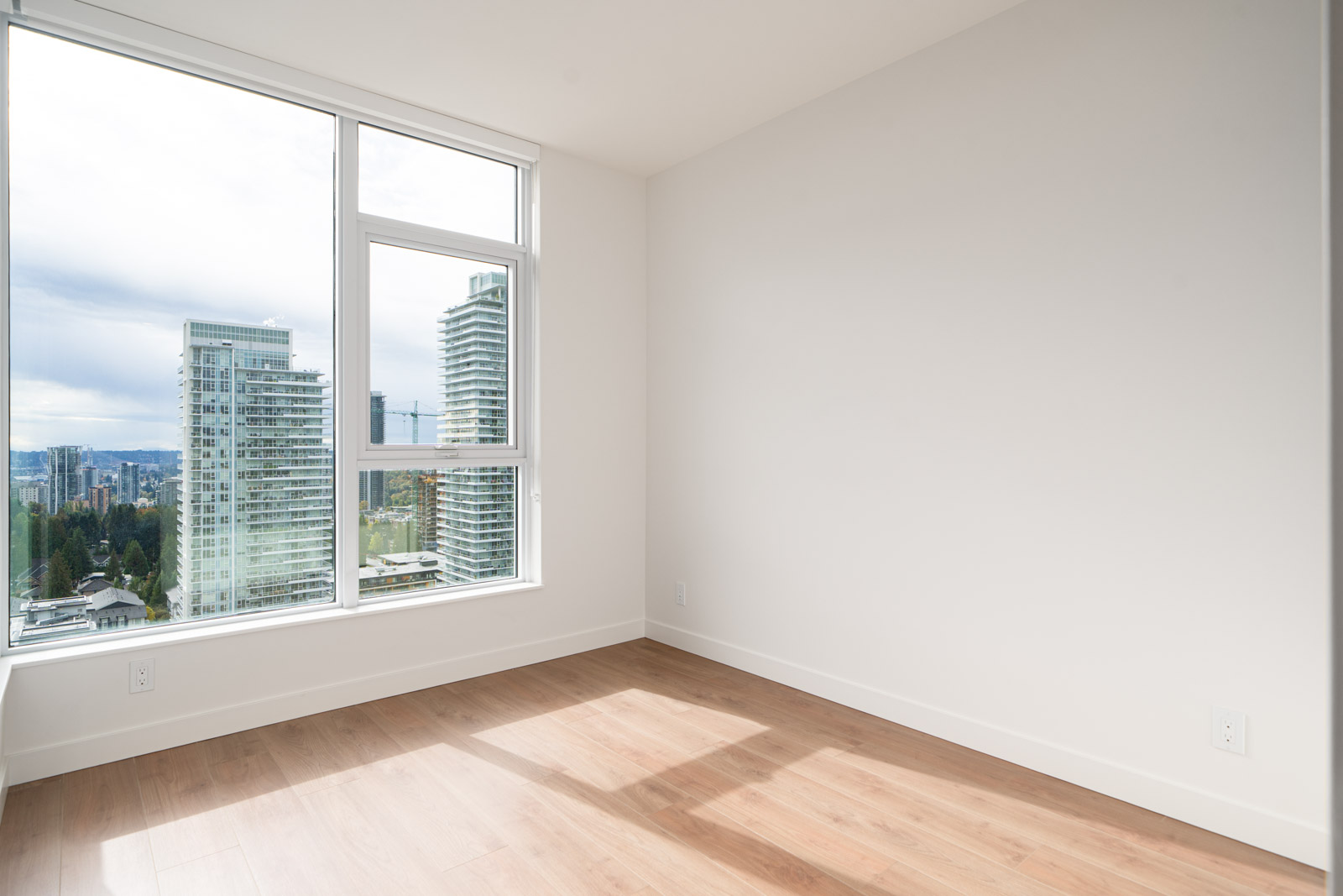 Empty, unfurnished room with light wood flooring, large window, and a view of city skyscrapers and cloudy sky outside. Walls and ceiling are white.