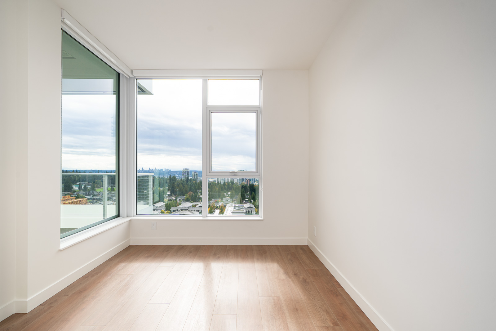 Empty room with light wood flooring, white walls, and large windows offering a view of a cityscape and cloudy sky.
