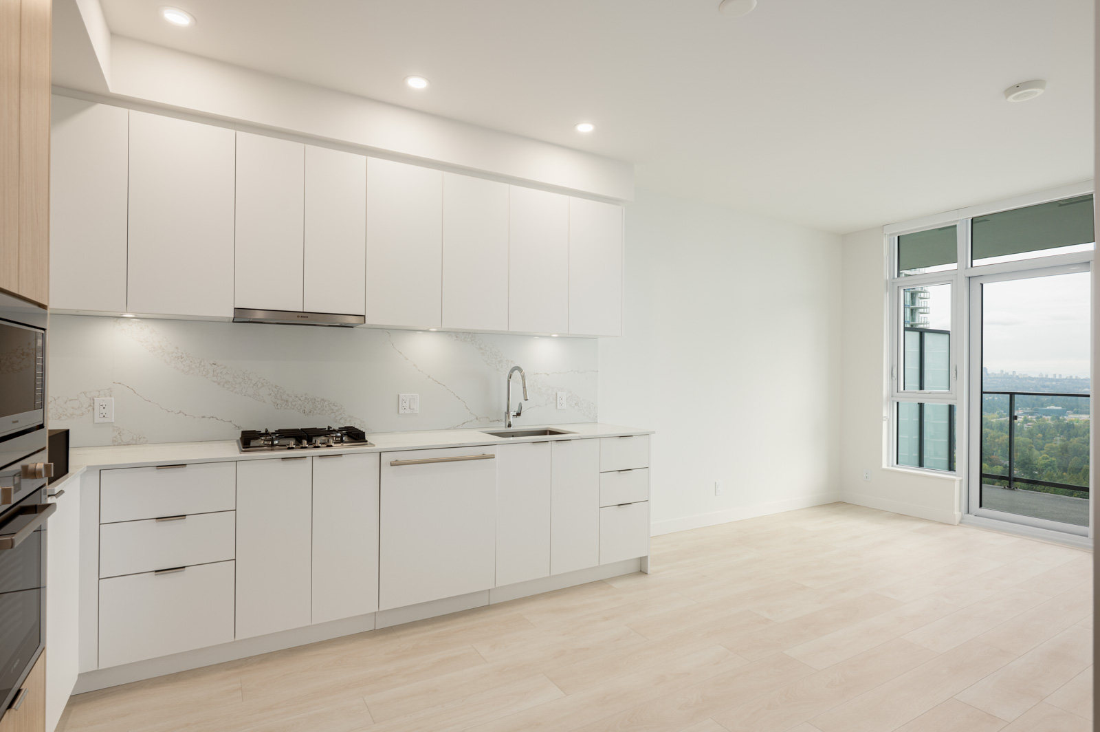 Modern kitchen with white cabinets, built-in appliances, light wood flooring, and a marble-patterned backsplash; large window with a view in the background.