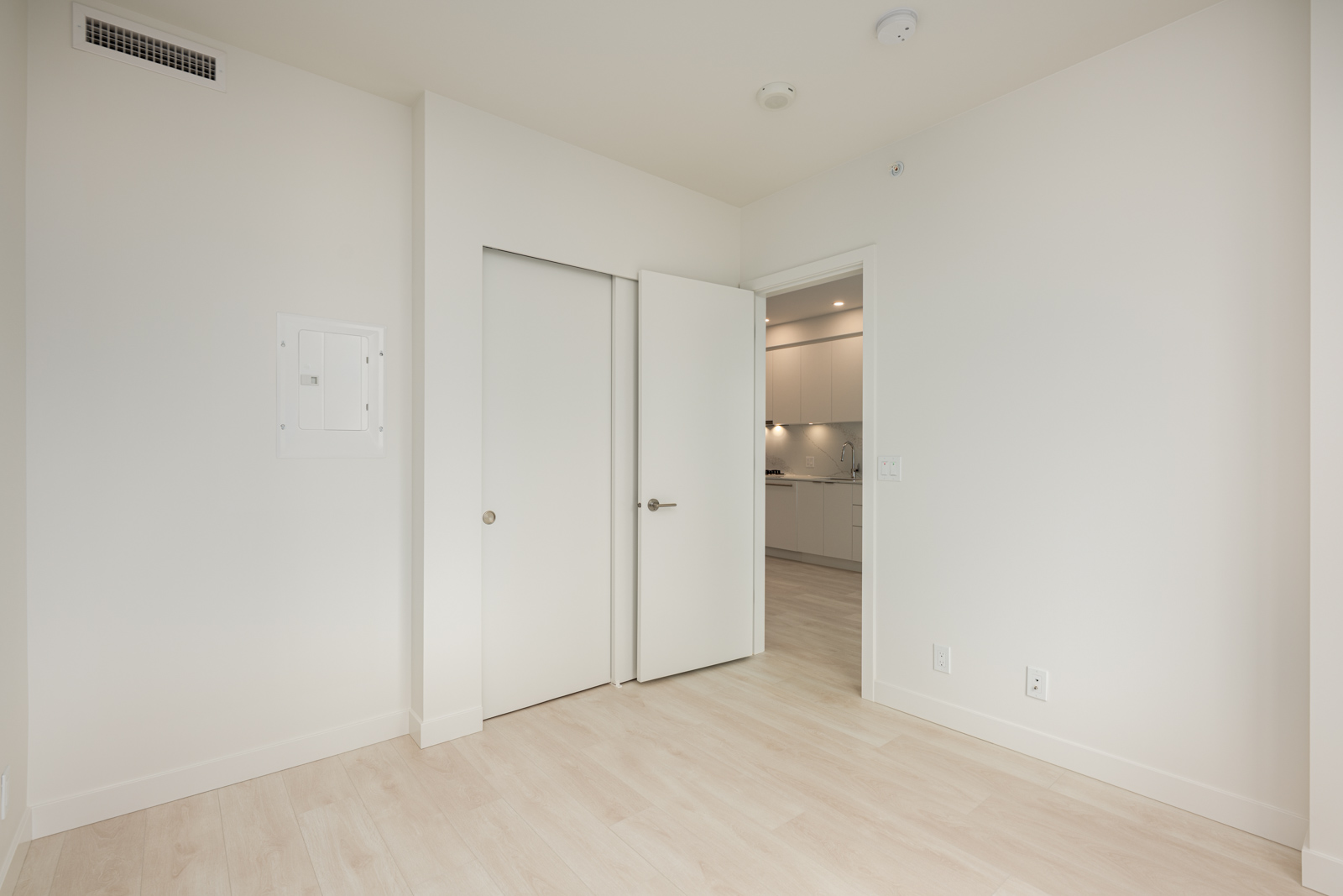 Empty room with light wood flooring, white walls, a closed closet door, an open door leading to a kitchen area, and a small vent on the upper wall.