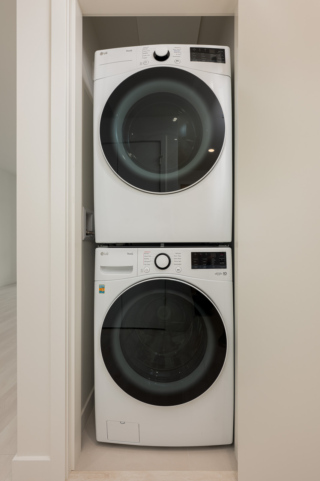 A stacked front-loading washer and dryer set is installed in a small, recessed laundry space with light-colored walls and flooring.