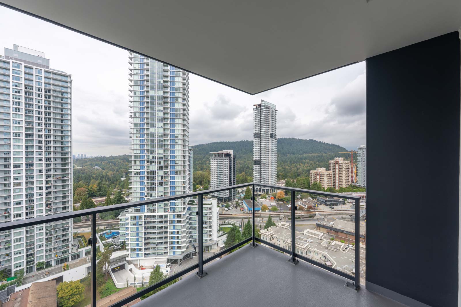 High-rise balcony with glass railing overlooking city buildings, roads, and a forested hillside under a cloudy sky.