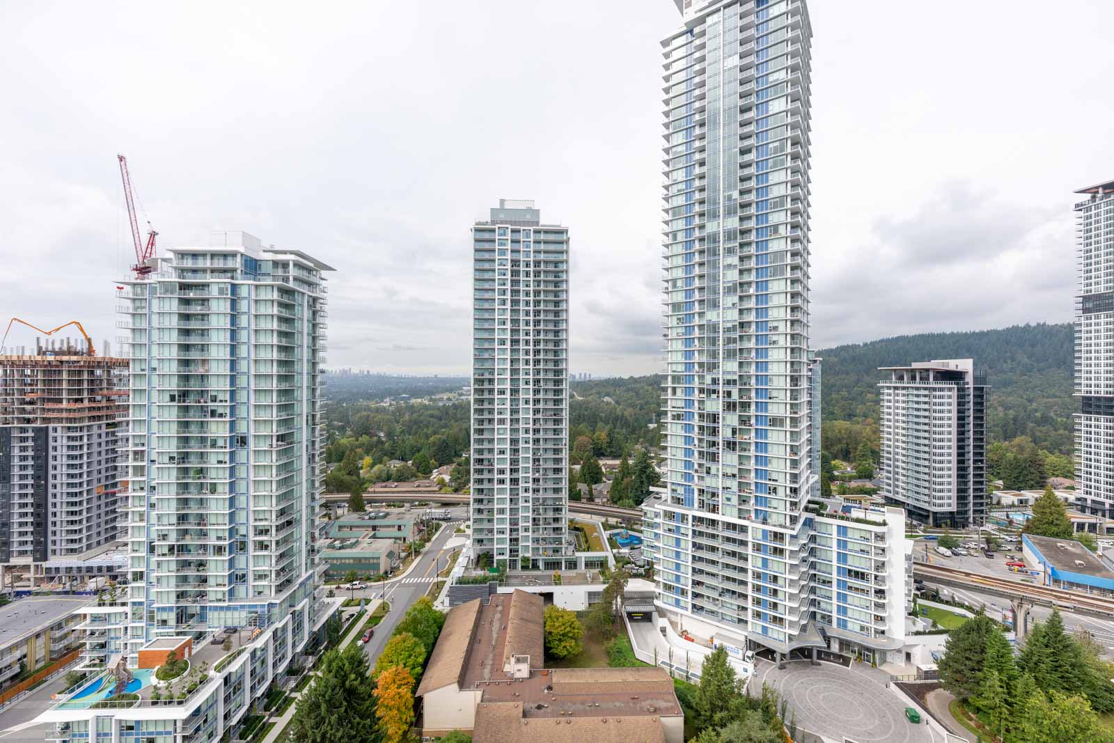 View of modern high-rise residential buildings with a construction crane and a forested area in the background under a cloudy sky.