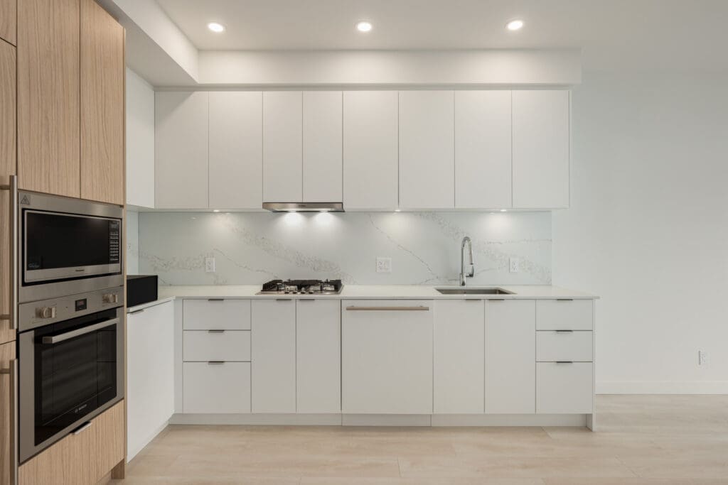 Modern kitchen with white cabinets, marble backsplash, built-in oven, stovetop, dishwasher, and light wood accents under recessed ceiling lights.