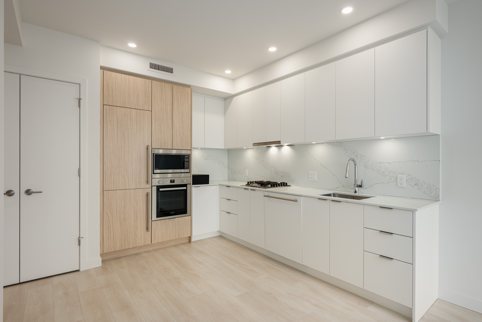 Modern kitchen with white cabinets, light wood accents, built-in oven and microwave, gas stove, under-cabinet lighting, and light wood flooring.