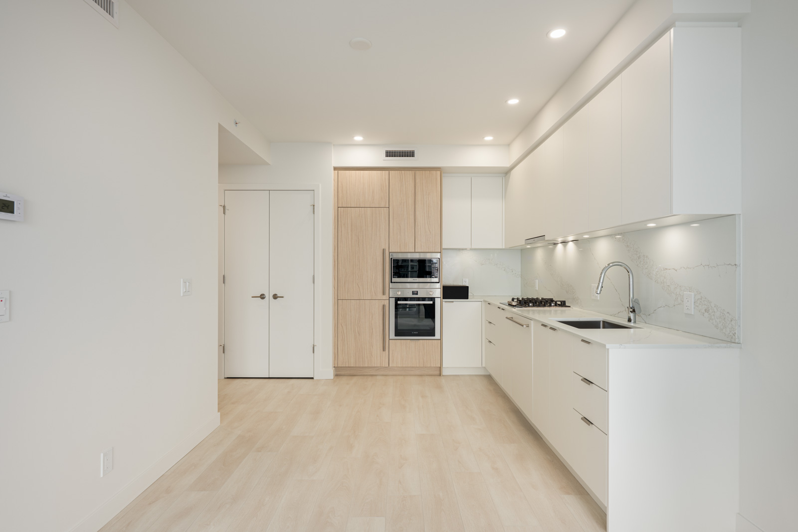 Modern kitchen with light wood floors, white cabinets, built-in oven, stovetop, and minimalist design. Recessed lighting and marble backsplash are visible.
