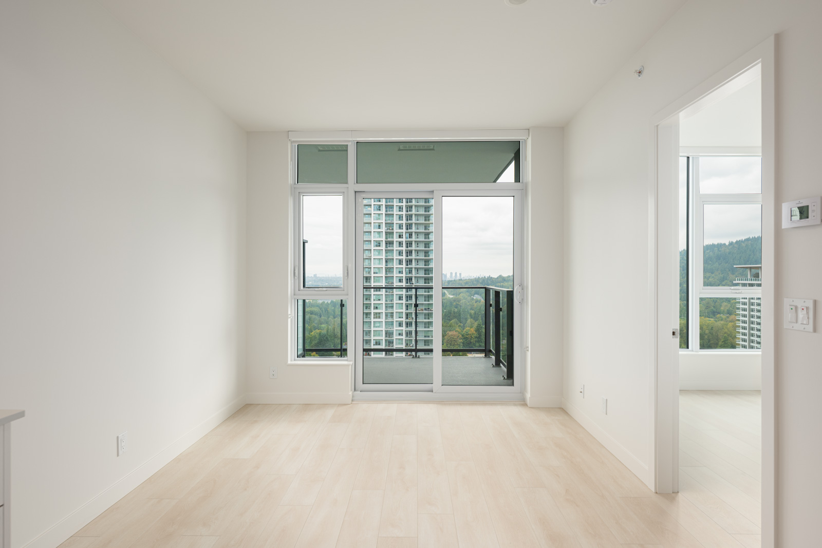 Empty, modern apartment room with light wood flooring, large windows, and a glass door leading to a balcony overlooking a cityscape with tall buildings and greenery.