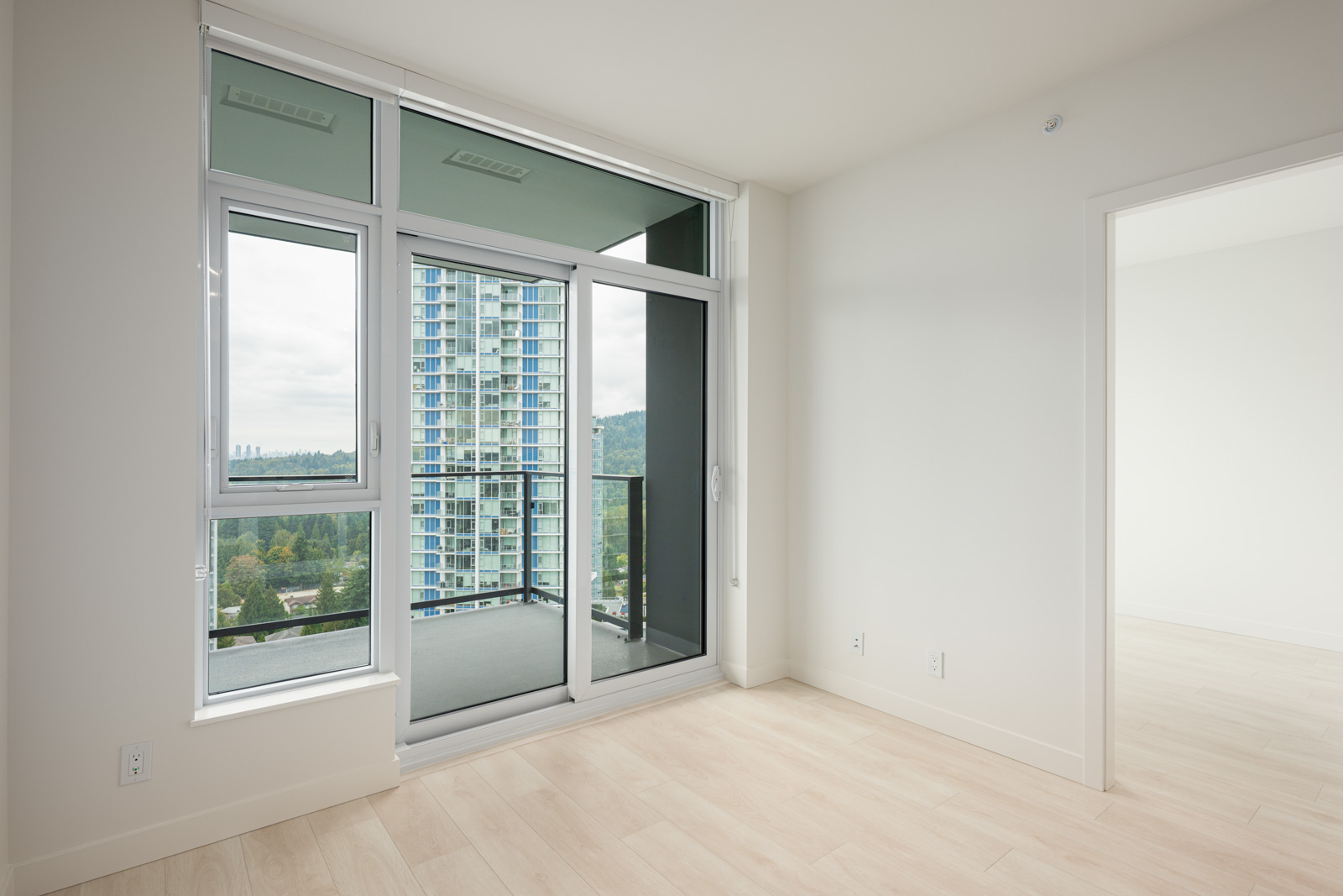 Empty modern apartment room with light wood floors, floor-to-ceiling windows, a balcony, and a view of a neighboring high-rise building.