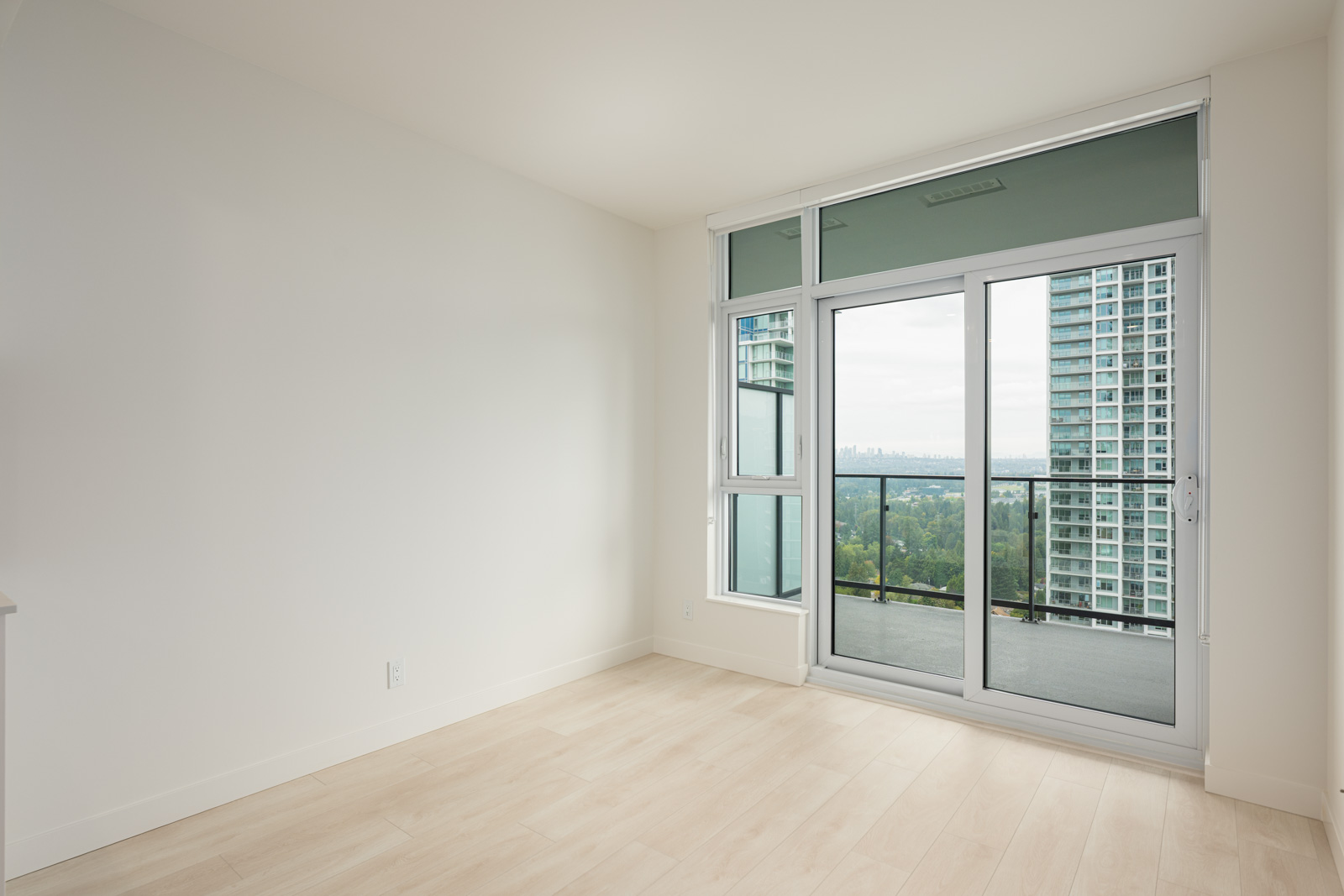 Empty room with light wood flooring, white walls, and large sliding glass doors leading to a balcony with city and greenery views.