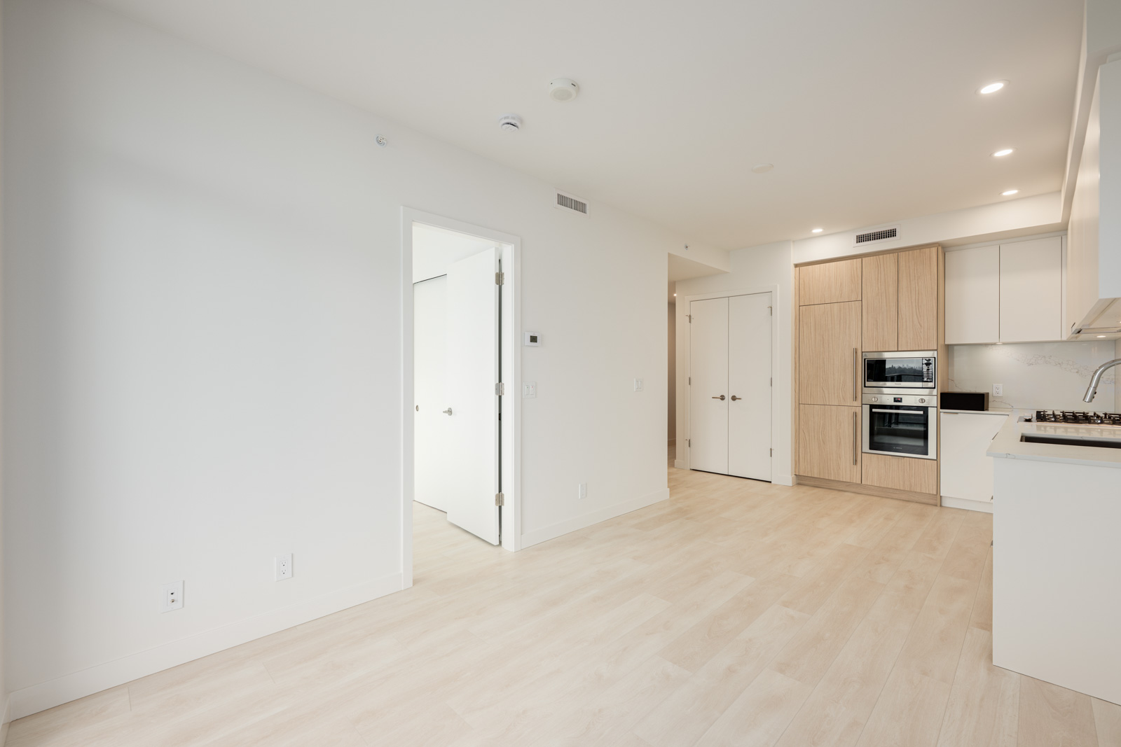 Modern, unfurnished apartment interior with light wood flooring, white walls, open kitchen area, built-in appliances, and a partially open door to another room.