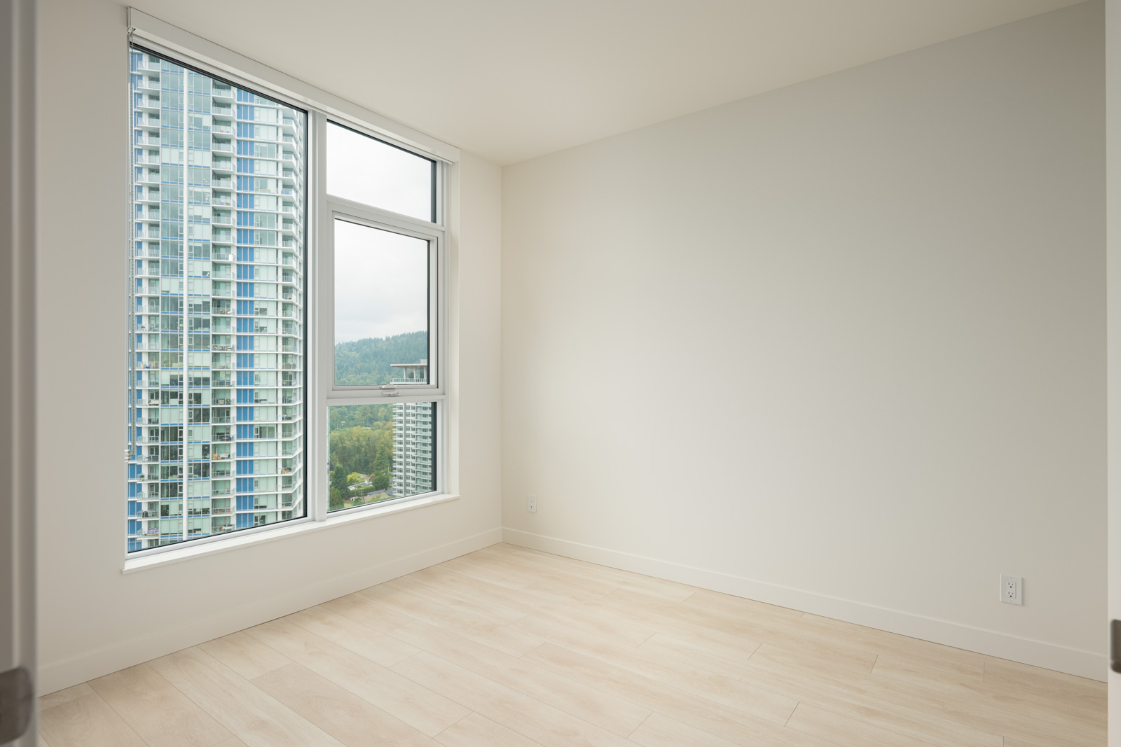 Empty room with light-colored wood flooring, white walls, and large corner windows showing a cityscape with tall buildings and greenery outside.