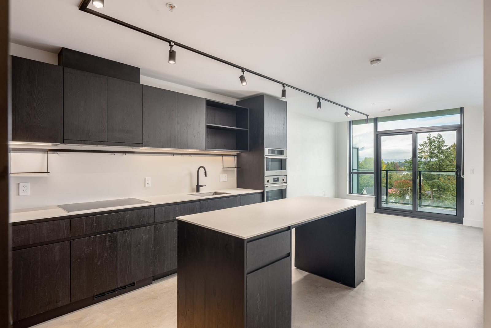 Modern kitchen with black cabinets, built-in appliances, island, and large windows overlooking a balcony with trees outside.