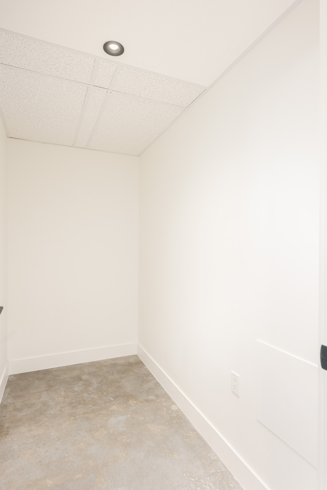 Small empty room with white walls, a gray concrete floor, white ceiling tiles, recessed lighting, and a visible power outlet on the right wall.