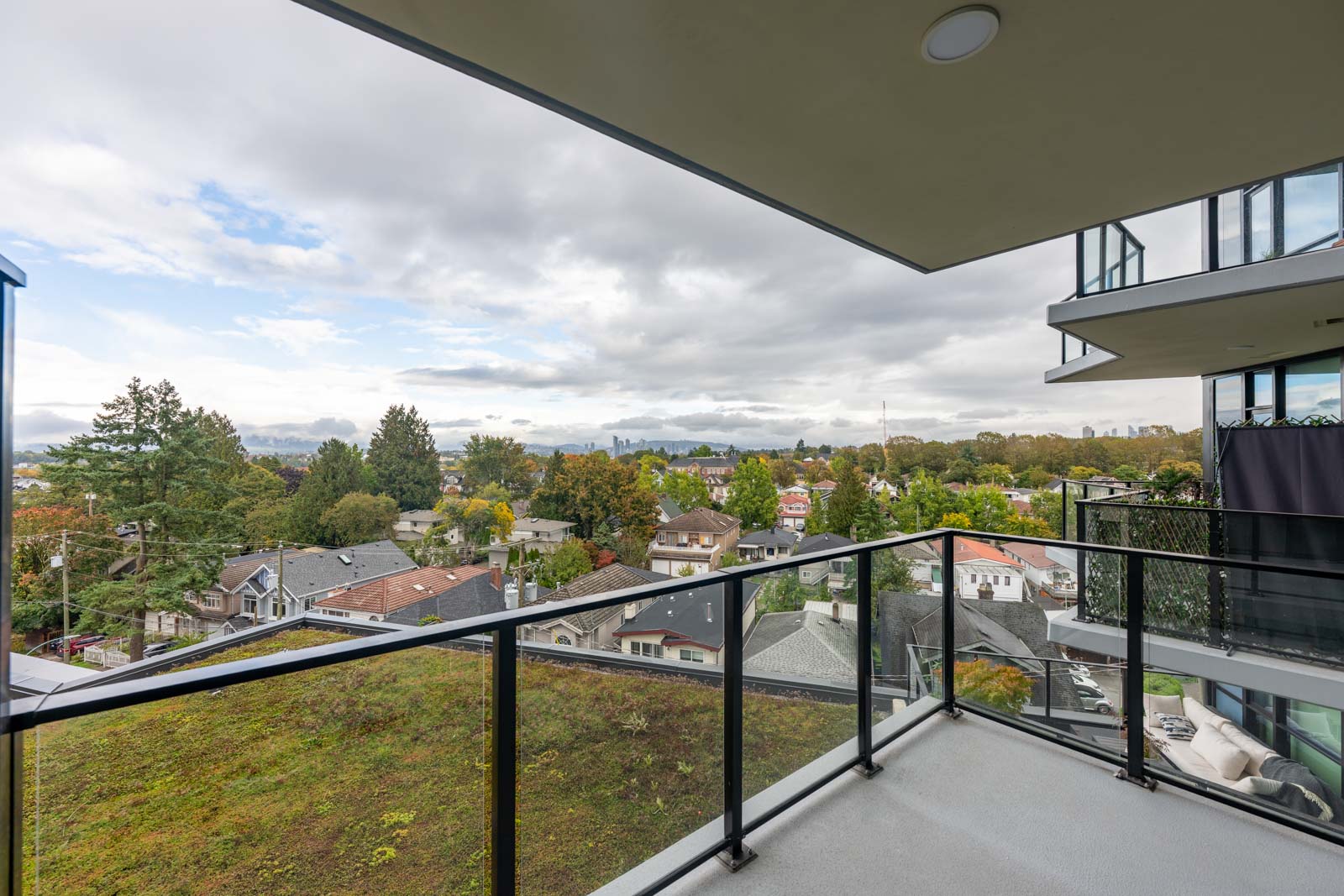 A glass balcony overlooks a residential neighborhood with houses, trees, and a cloudy sky in the background.