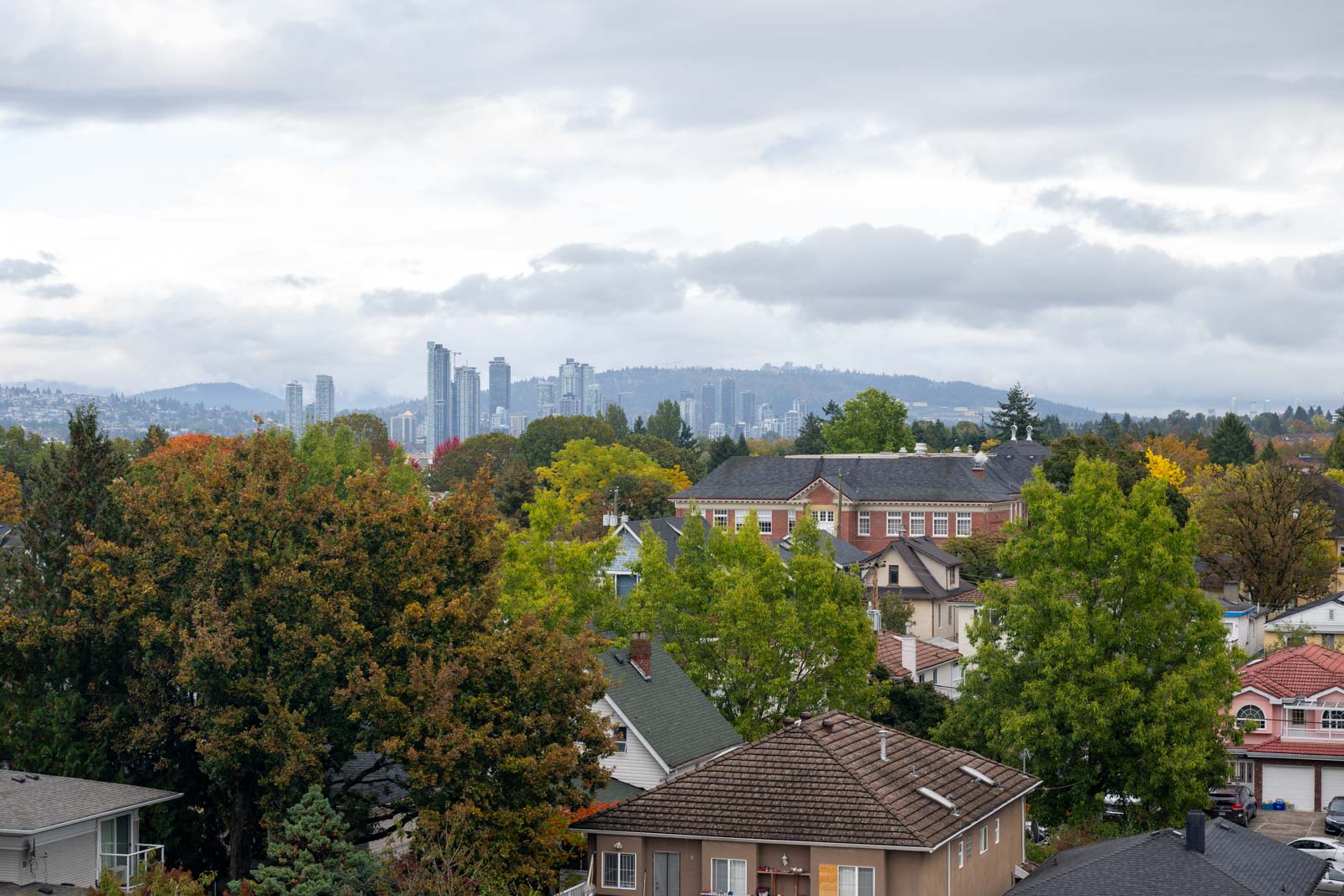 View of suburban houses with trees in the foreground and a city skyline with high-rise buildings and hills in the background under a cloudy sky.