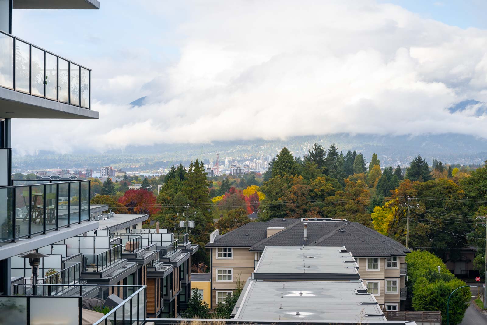 View of residential buildings and trees with autumn foliage, under a cloudy sky partially covering distant mountains.