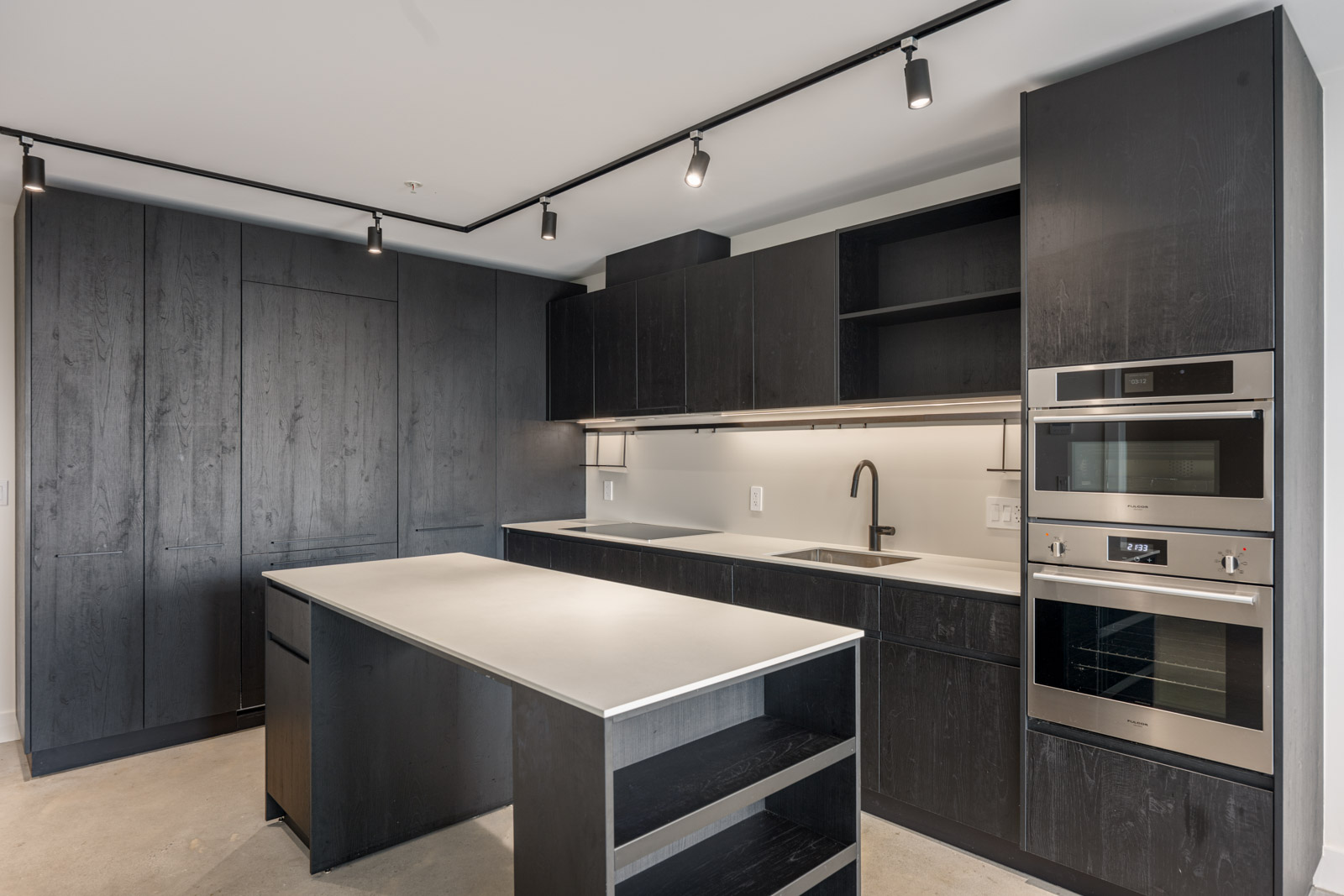 Modern kitchen with dark cabinets, built-in appliances, a light countertop island, open shelving, and track lighting on the ceiling.