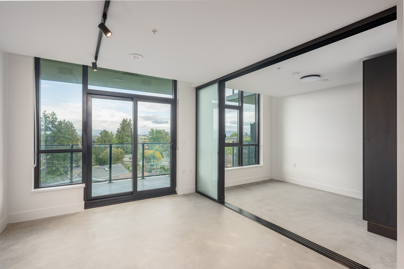 A modern, unfurnished apartment room with large windows, glass sliding doors, and concrete floors, overlooking a green landscape.
