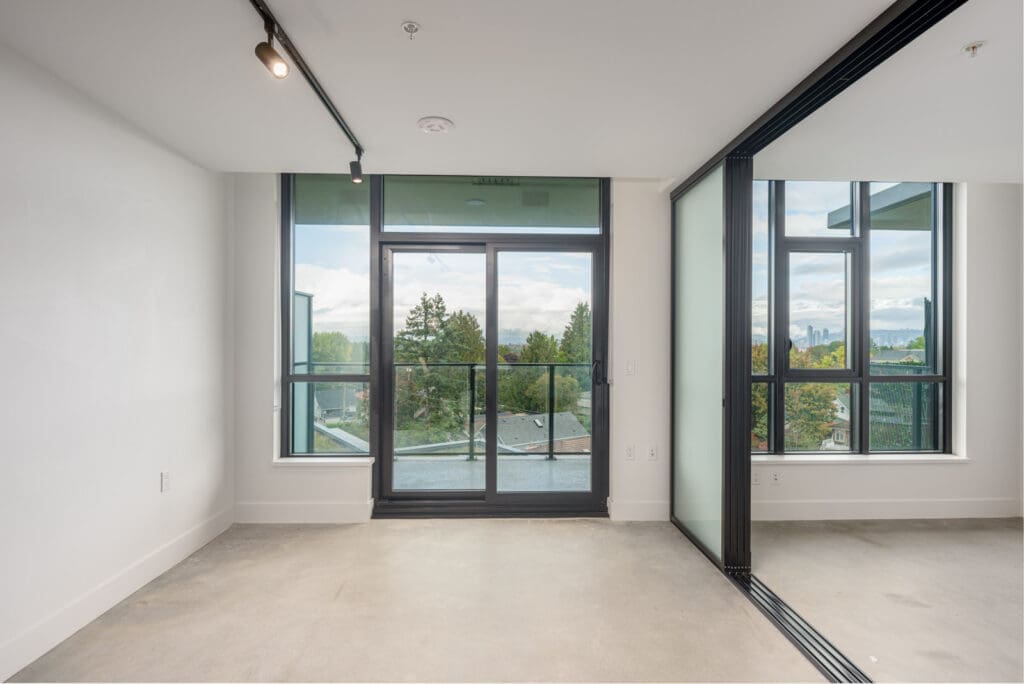 An empty modern room with large windows and a glass door leading to a balcony, offering a view of trees and distant buildings under a cloudy sky.