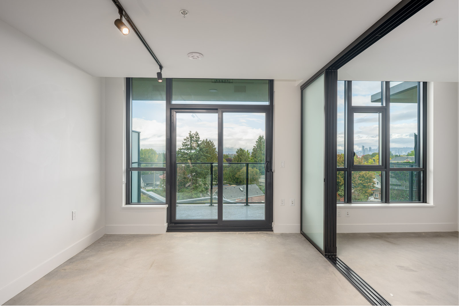 An empty modern room with large windows and a glass door leading to a balcony, offering a view of trees and distant buildings under a cloudy sky.