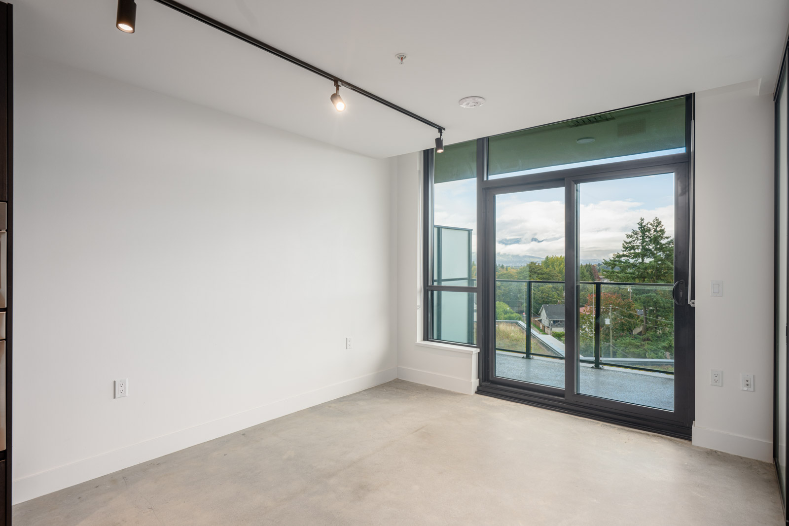 Empty room with white walls, concrete floor, large glass doors, and a balcony overlooking trees and a cloudy sky. Ceiling track lighting is visible.