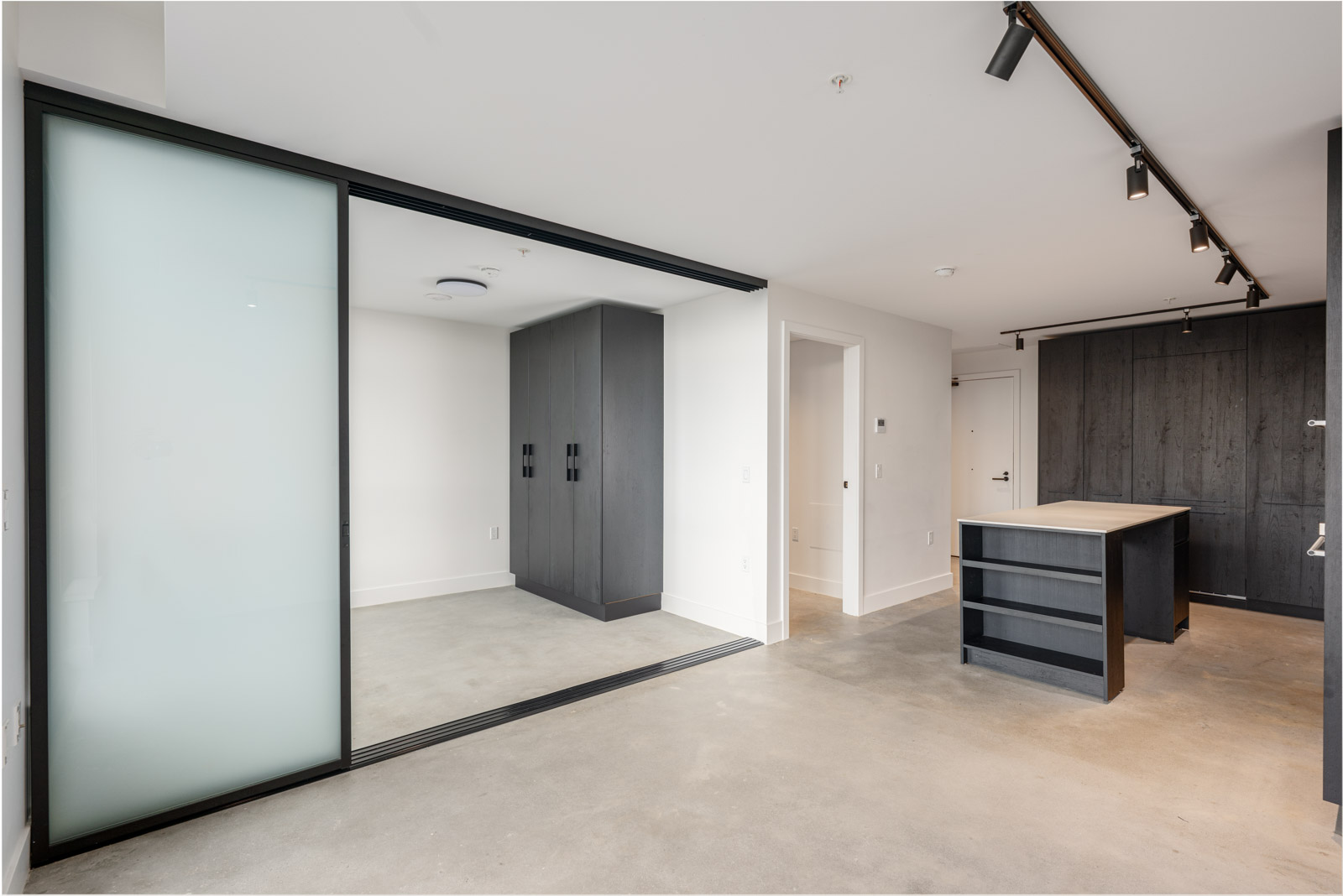 Modern, unfurnished apartment interior featuring concrete floors, a kitchen with dark cabinetry, an island, and a frosted glass sliding door separating a small room or area.