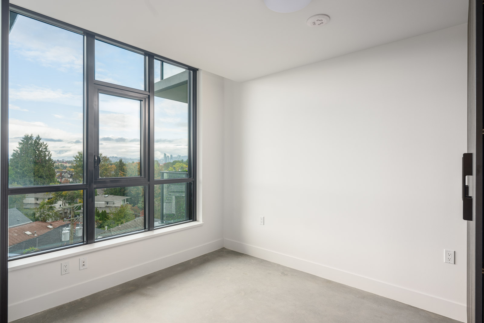Empty room with white walls, large floor-to-ceiling windows, and a concrete floor, overlooking a neighborhood with trees and houses outside.